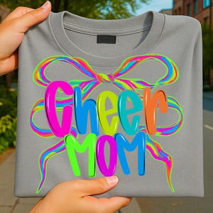 May include: A gray sweatshirt with the words "Cheer MOM" in bright, colorful letters. The text is overlaid on a neon rainbow bow design. The sweatshirt is held by hands.
