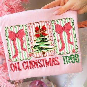 May include: A light pink t-shirt featuring three framed designs. The designs include a Christmas tree with a red bow, and two pink bows with green and white striped backgrounds. The text "OH. CHRISTMAS TREE" is printed below the designs.