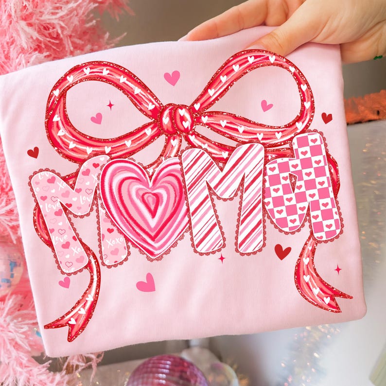 May include: A light pink t-shirt with a large red bow and the word "MOM" in a decorative font. The "O" is a heart-shaped design. Small hearts and stars are scattered around the design. The shirt is held by a hand.