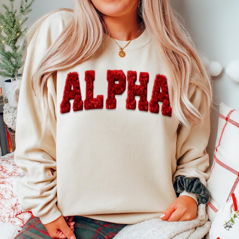 May include: Cream-colored sweatshirt with the word "ALPHA" in large, textured, red letters across the chest. The sweatshirt has a crew neck and long sleeves. The model is wearing a gold necklace.