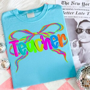 Op de afbeelding: Een lichtblauw t-shirt met het woord "Teacher" in kleurrijke bubbelletters. Het woord is versierd met een regenboogkleurig strikdesign. Het shirt wordt gepresenteerd op een roze en wit geblokt oppervlak.
