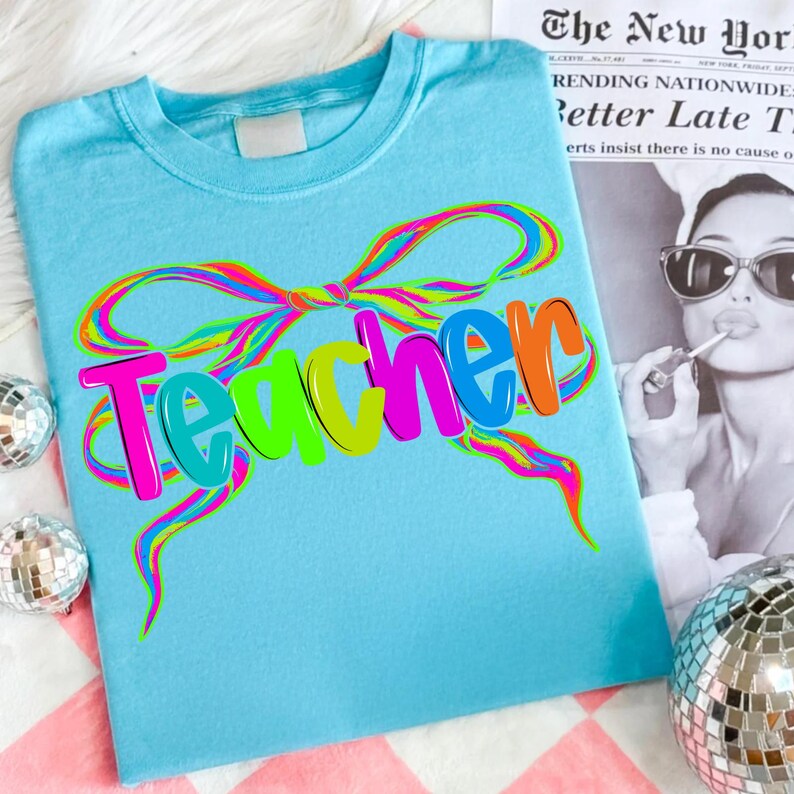 Op de afbeelding: Een lichtblauw t-shirt met het woord "Teacher" in kleurrijke bubbelletters. Het woord is versierd met een regenboogkleurig strikdesign. Het shirt wordt gepresenteerd op een roze en wit geblokt oppervlak.