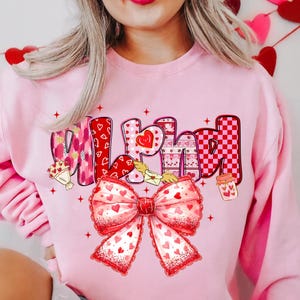 May include: Pink sweatshirt with the word "Wifey" in a Valentine's Day theme. The letters are decorated with hearts, patterns, and a large bow. A bouquet of flowers and a jar of hearts are also featured.