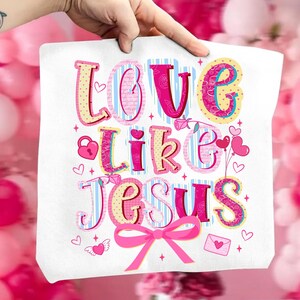 May include: White tote bag with the words "Love Like Jesus" in a colorful, whimsical font. The design includes hearts, a lock, a bow, and a small envelope. The background is a soft pink.
