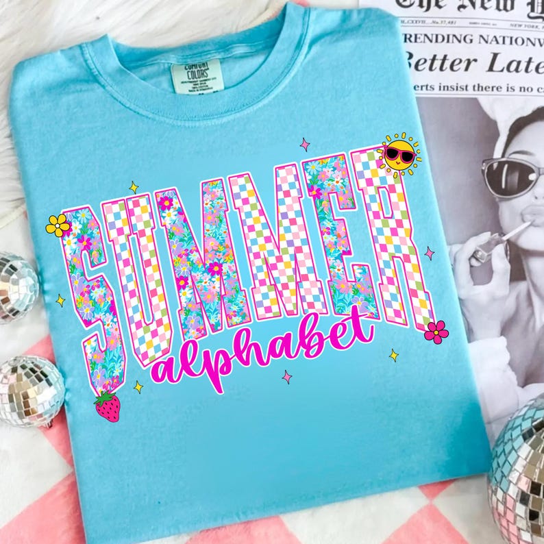 May include: A light blue t-shirt with the words "SUMMER alphabet" in a floral and checkered design. The shirt features a cartoon sun wearing sunglasses, a strawberry, and small flower and star graphics.