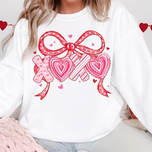 May include: White sweatshirt with a large red bow and the letters "XOX" with heart designs. The bow and letters are decorated with pink and red hearts. Small pink hearts are scattered around the design. A Valentine's Day themed top.