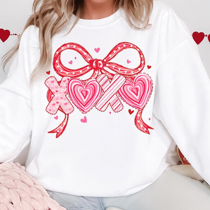 May include: White sweatshirt with a large red bow and the letters "XOX" with heart designs. The bow and letters are decorated with pink and red hearts. Small pink hearts are scattered around the design. A Valentine's Day themed top.
