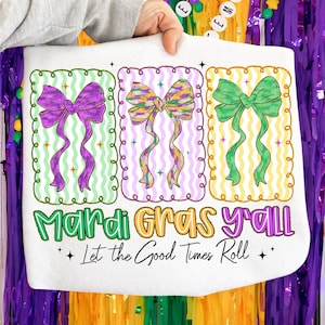 May include: White tote bag featuring three colorful bow designs in purple, gold, and green, each within a decorative frame. The text "Mardi Gras y'all" and "Let the Good Times Roll" are printed below the bows, celebrating the festive season.