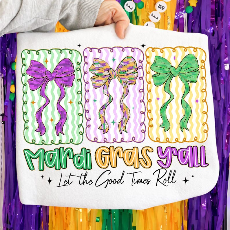 May include: White tote bag featuring three colorful bow designs in purple, gold, and green, each within a decorative frame. The text "Mardi Gras y'all" and "Let the Good Times Roll" are printed below the bows, celebrating the festive season.