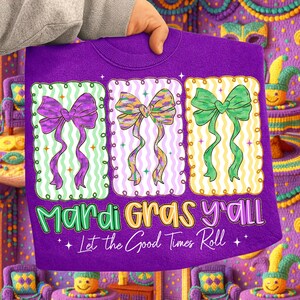 May include: A purple sweatshirt featuring three decorative bows in purple, yellow, and green, each set within a rectangular frame. The text "Mardi Gras y'all" and "Let the Good Times Roll" are printed below the bows. The sweatshirt is held by a person.