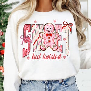 May include: White sweatshirt featuring the words "SWEET" and "but twisted" in red and pink. A pink gingerbread man with a red bow tie is in the center. The design includes snowflakes and bows.