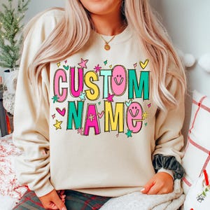 May include: A beige sweatshirt with the words "CUSTOM NAME" in colorful, bubble letters. The design includes stars, hearts, and smiley faces. The sweatshirt is a casual, comfortable style.