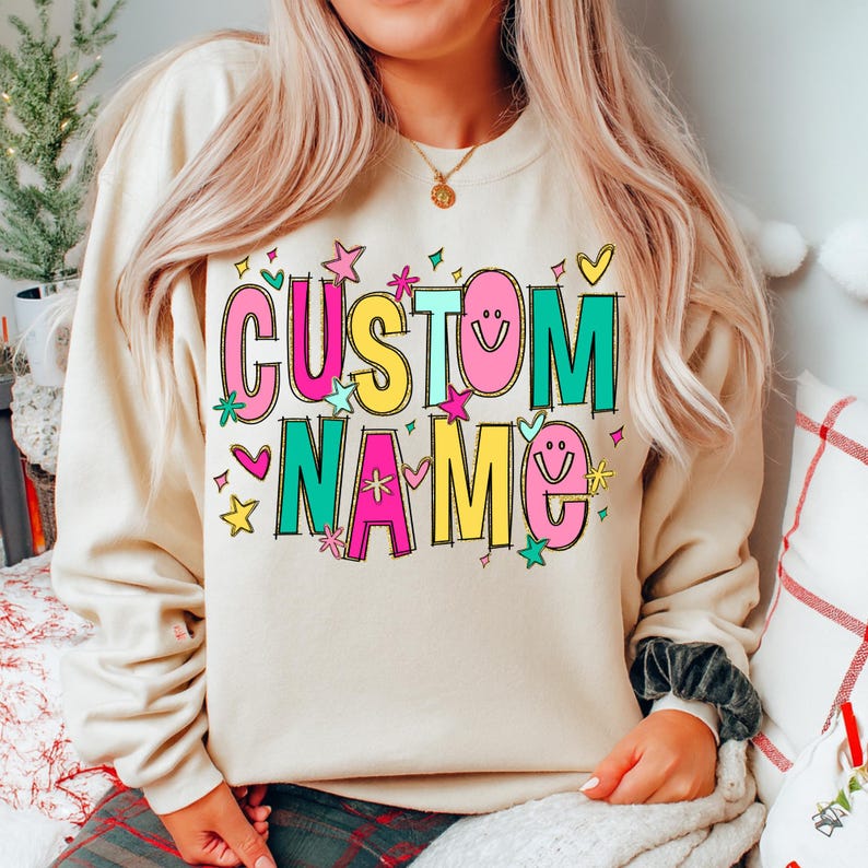 May include: A beige sweatshirt with the words "CUSTOM NAME" in colorful, bubble letters. The design includes stars, hearts, and smiley faces. The sweatshirt is a casual, comfortable style.