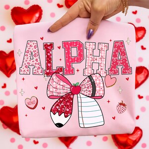 Op de afbeelding: Roze sweatshirt met het woord "ALPHA" in verschillende patronen en kleuren. Het ontwerp bevat een grote strik met een potlood, appel en notitieboek, plus harten en aardbeien. Het sweatshirt wordt door een hand omhoog gehouden.
