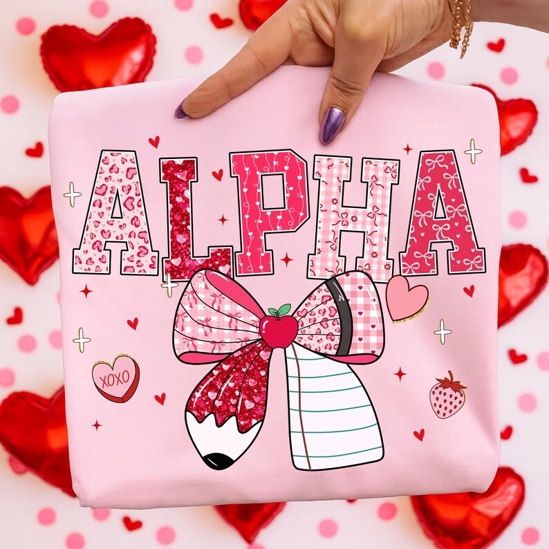 Op de afbeelding: Roze sweatshirt met het woord "ALPHA" in verschillende patronen en kleuren. Het ontwerp bevat een grote strik met een potlood, appel en notitieboek, plus harten en aardbeien. Het sweatshirt wordt door een hand omhoog gehouden.