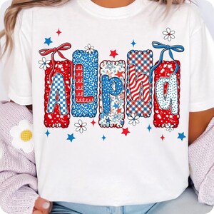 May include: White t-shirt with the word "ALPHA" in red, white, and blue patterns. The letters are decorated with stars, checks, and floral accents. The design is surrounded by small stars and flowers.