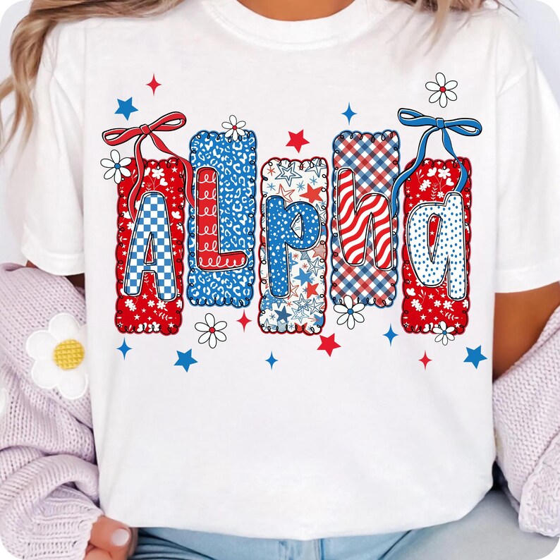 May include: White t-shirt with the word "ALPHA" in red, white, and blue patterns. The letters are decorated with stars, checks, and floral accents. The design is surrounded by small stars and flowers.