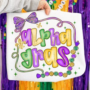 May include: White tote bag with the words "Alpha Gras" in purple, green, and gold lettering. The design includes a purple bow, a fleur-de-lis, and a beaded necklace. The bag is held up by a person.