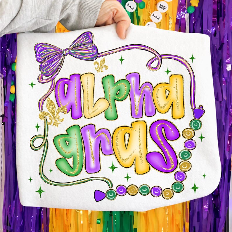 May include: White tote bag with the words "Alpha Gras" in purple, green, and gold lettering. The design includes a purple bow, a fleur-de-lis, and a beaded necklace. The bag is held up by a person.