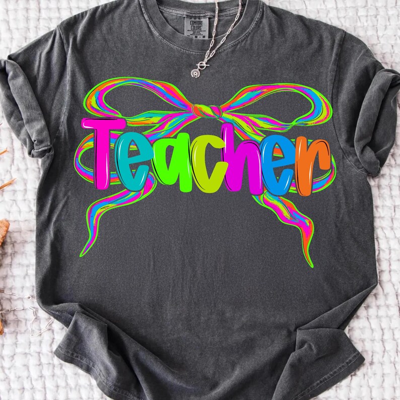 Op de afbeelding: Donkergrijs t-shirt met het woord "Teacher" in kleurrijke, bubbelletters. Het woord wordt omlijst door een levendig, regenboogkleurig strikdesign. Een zilveren ketting is zichtbaar aan de bovenkant.