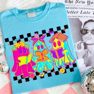 May include: A light blue t-shirt featuring a colorful graphic of three ghost-like figures reading books. The figures are in pink, blue, and yellow with floral and star accents. A black and white checkered pattern borders the design.