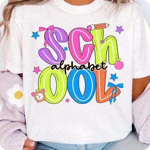 May include: White t-shirt featuring a vibrant graphic. The word "SCHOOL" is displayed in large, outlined letters with "alphabet" underneath. The design includes a pencil, apple, stars, and bows, perfect for back to school.