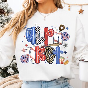 Op de afbeelding: Witte sweatshirt met de tekst "All About" in een honkbalthema. De letters zijn rood, blauw en zwart-wit geruit, met honkballen, knuppels en sterren. Een leuk en sportief ontwerp.