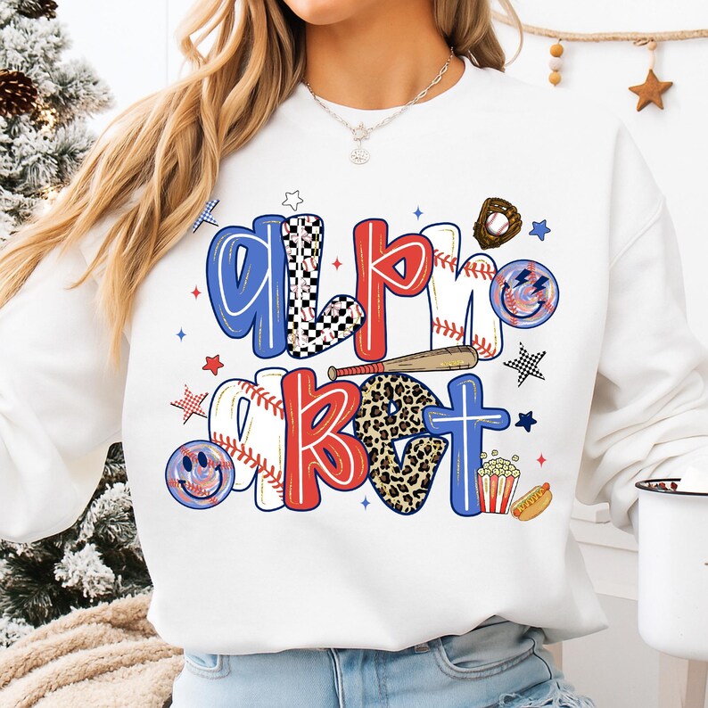 Op de afbeelding: Witte sweatshirt met de tekst "All About" in een honkbalthema. De letters zijn rood, blauw en zwart-wit geruit, met honkballen, knuppels en sterren. Een leuk en sportief ontwerp.