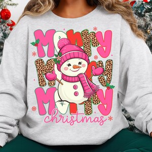 May include: A light gray sweatshirt featuring a snowman graphic with a pink hat, scarf, and mittens. The words "Merry Christmas" are displayed in pink and leopard print lettering, with holly accents. A festive holiday design.