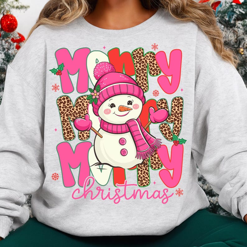 May include: A light gray sweatshirt featuring a snowman graphic with a pink hat, scarf, and mittens. The words "Merry Christmas" are displayed in pink and leopard print lettering, with holly accents. A festive holiday design.