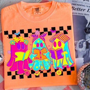 May include: Orange Comfort Colors t-shirt featuring a graphic of three cartoon ghosts reading books. The ghosts are in bright neon colors with floral and star accents. A black and white checkered pattern and bats are above the ghosts.