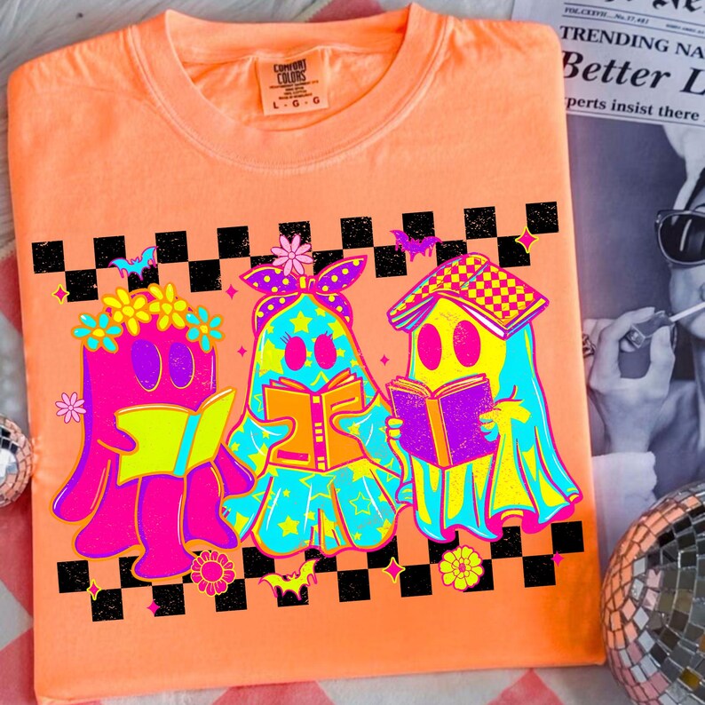 May include: Orange Comfort Colors t-shirt featuring a graphic of three cartoon ghosts reading books. The ghosts are in bright neon colors with floral and star accents. A black and white checkered pattern and bats are above the ghosts.