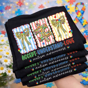 May include: Black t-shirts with the words "ACCEPT-UNDERSTAND-LOVE" and "autism awareness" printed on them. Each shirt features a colourful puzzle piece and a bow design. The shirts are stacked, with the text repeated.
