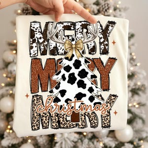 May include: Cream-colored t-shirt with the words "MERRY CHRISTMAS MERRY" in a mix of brown cow print, leopard print, and glittery brown lettering. A Christmas tree with a cow print design and antlers is in the center.