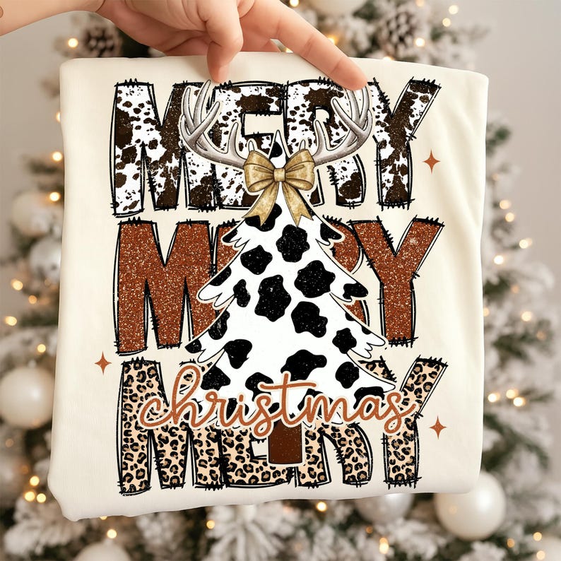 May include: Cream-colored t-shirt with the words "MERRY CHRISTMAS MERRY" in a mix of brown cow print, leopard print, and glittery brown lettering. A Christmas tree with a cow print design and antlers is in the center.