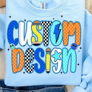 May include: Light blue sweatshirt with the words "CUSTOM DESIGN" in a colorful, cartoonish font. The design includes a pencil, lightning bolt, star, and smiley faces. The sweatshirt is made of a soft material.