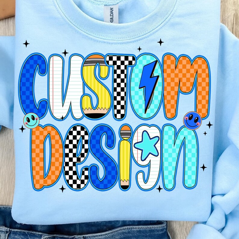 May include: Light blue sweatshirt with the words "CUSTOM DESIGN" in a colorful, cartoonish font. The design includes a pencil, lightning bolt, star, and smiley faces. The sweatshirt is made of a soft material.