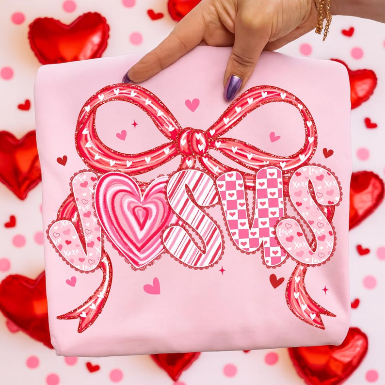 May include: Pink sweatshirt with a large bow and the word "JESUS" in a heart-themed design. The letters are decorated with hearts and patterns. The sweatshirt is held up by a hand.