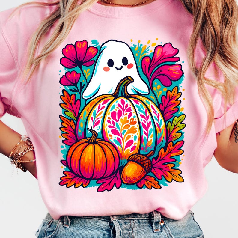 May include: Pink t-shirt featuring a colorful Halloween design. A smiling white ghost hovers above a large pumpkin with a floral pattern. An orange pumpkin and acorn are below, surrounded by pink and teal foliage.