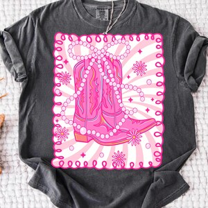 May include: Dark gray t-shirt featuring a pink graphic of cowboy boots adorned with a pearl necklace. The boots are set against a pink and white striped background with floral accents, all within a decorative pink border.