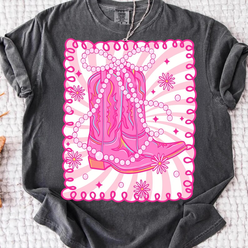 May include: Dark gray t-shirt featuring a pink graphic of cowboy boots adorned with a pearl necklace. The boots are set against a pink and white striped background with floral accents, all within a decorative pink border.