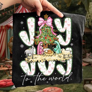 May include: Black t-shirt with a Christmas Nativity scene. The word "JOY" is in large white letters with a pink bow and Christmas tree. The text "to the world" is written below the Nativity scene.