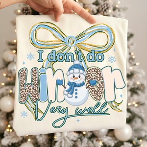May include: Cream-colored t-shirt with a whimsical winter design. The shirt features a snowman wearing a blue hat and scarf, with the text "I don't do winter very well." The word "winter" has a leopard print pattern. A blue and gold bow is at the top.