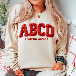 May include: A cream-colored sweatshirt with the letters "ABCD" in a bold, red, knitted font. Below the letters, the text reads "KNITTED ALPHA" with red heart symbols. The sweatshirt is a casual, comfortable style.