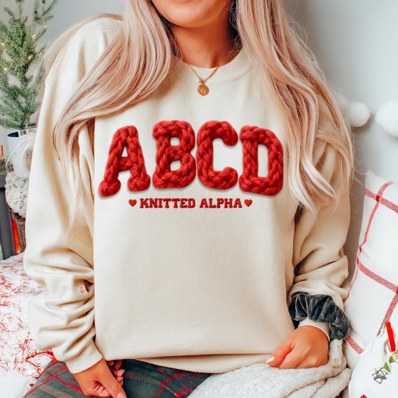 May include: A cream-colored sweatshirt with the letters "ABCD" in a bold, red, knitted font. Below the letters, the text reads "KNITTED ALPHA" with red heart symbols. The sweatshirt is a casual, comfortable style.