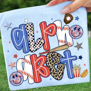Op de afbeelding: Lichtblauw T-shirt met de woorden "ALMOST BASEBALL" in rood, wit, blauw en luipaardprint. Honkbalthema-afbeeldingen zijn onder meer een knuppel, bal, handschoen, sterren en een smiley.