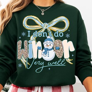 May include: Dark green sweatshirt with the text "I don't do winter very well." The design includes a snowman wearing a blue hat and scarf, and the word "winter" has a leopard print pattern. A large blue and gold bow is at the top.