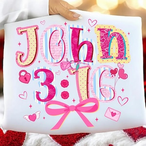 May include: White cushion featuring the text "John 3:16" in pink and yellow lettering. The design includes hearts, a bow, and a rose. The cushion is held by a person.