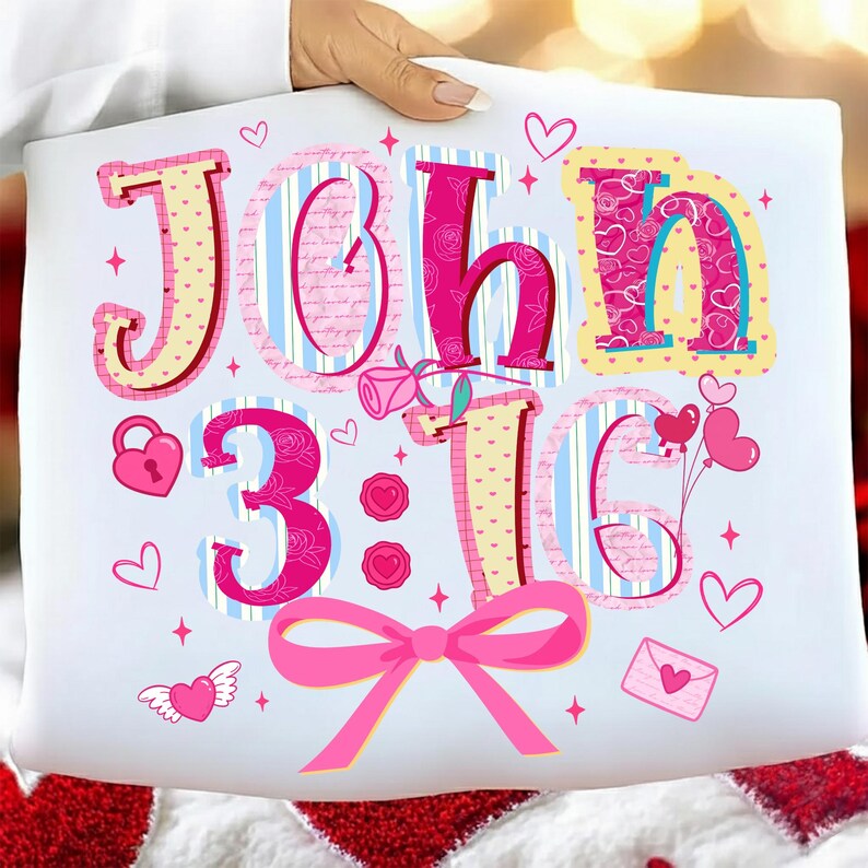 May include: White cushion featuring the text "John 3:16" in pink and yellow lettering. The design includes hearts, a bow, and a rose. The cushion is held by a person.
