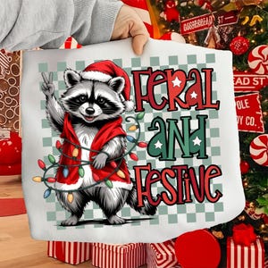 May include: White tote bag featuring a raccoon wearing a Santa hat and red jacket, holding a string of Christmas lights. The text "FERAL AND FESTIVE" is displayed in red and green.
